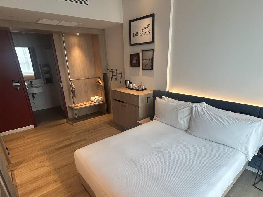 A modern hotel room with a large bed, wooden flooring, a small kitchenette, and a bathroom visible through an open door.