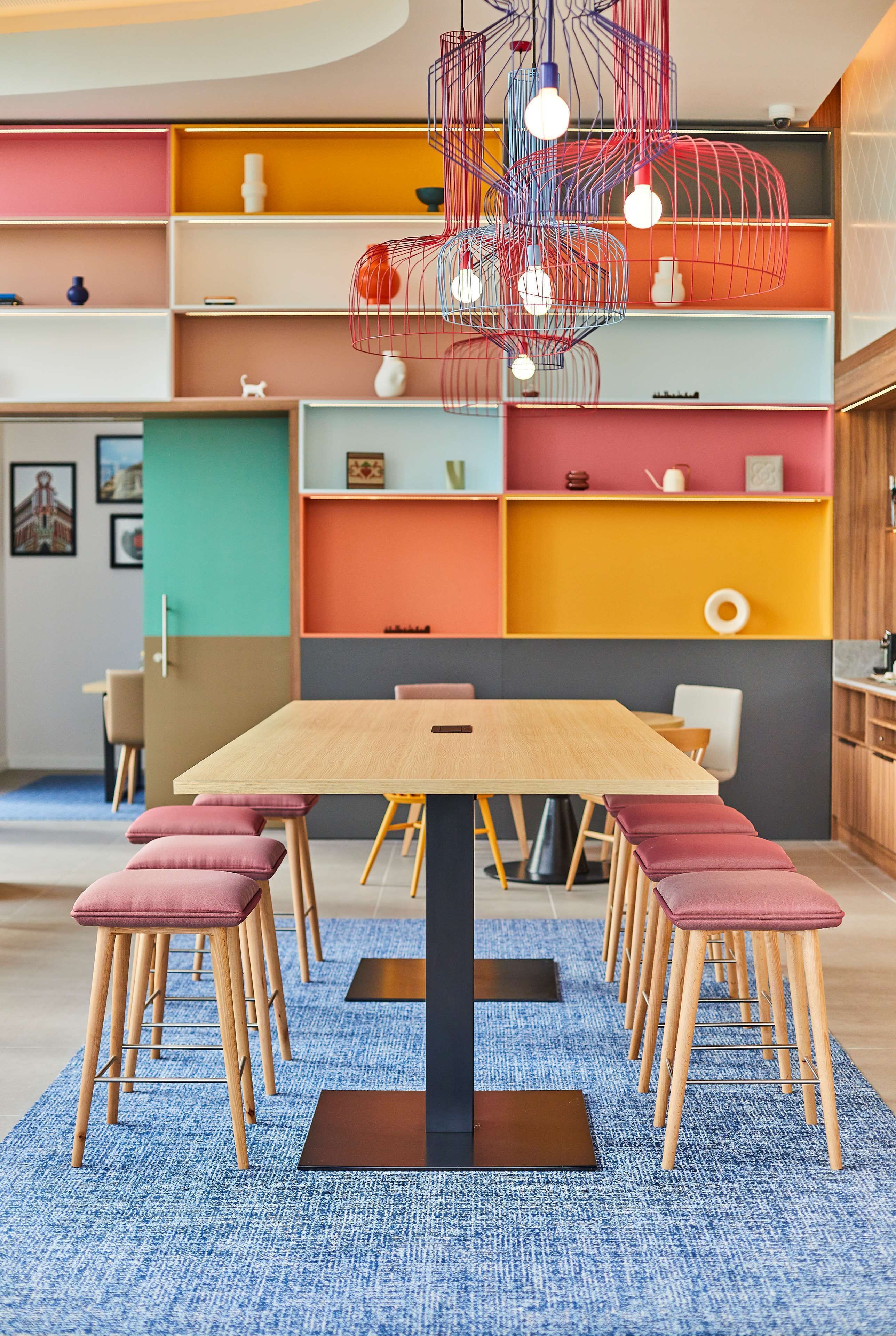 A modern dining area with a wooden table and matching chairs, a blue carpet, and colorful shelves in the background.