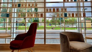 Un interior moderno con un ventanal que muestra una vista de un parque verde y edificios. Hay dos sillas, una roja y una marrón, y un separador de madera con letras que forman un mensaje.