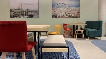Un comedor con una silla roja, un banco y una mesa con cuatro sillas. Hay dos cuadros en la pared que representan un paisaje urbano y una noria.