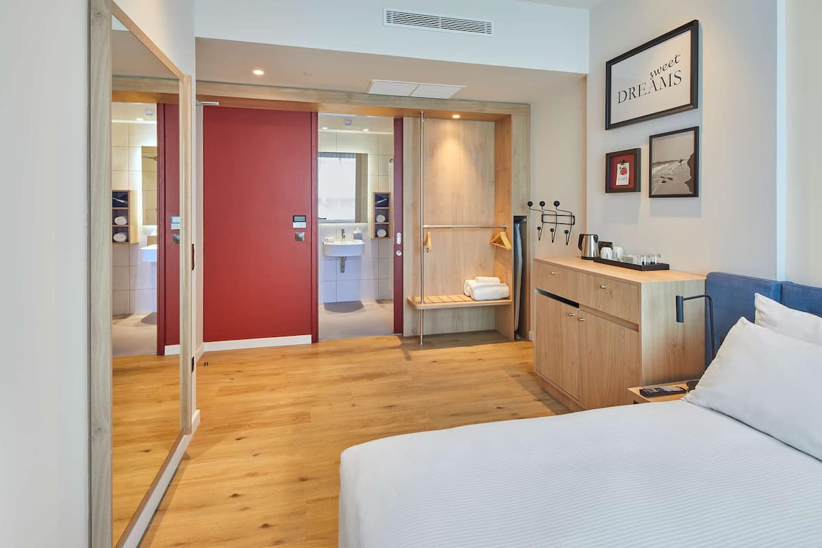 Een moderne hotelkamer met een houten vloer, een bed met witte lakens, een rode deur naar een badkamer en een wandplank met toiletartikelen.