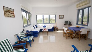A living room with a sofa, chairs, a coffee table, a TV, and large windows. - Family Holidays House Koutsouras by GHH (Ierapetra)