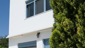 A modern white building with two windows and a door, a small tree in front, and two wooden lounge chairs. - Family Holidays House Koutsouras by GHH (Ierapetra)