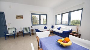 A bright living room with a blue sofa, striped chairs, a dining table with a fruit bowl, and large windows. - Family Holidays House Koutsouras by GHH (Ierapetra)