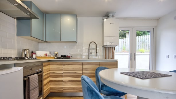 A modern kitchen with wooden cabinets, a white countertop, and a dining area with blue chairs. - Charming 1-bed Cottage in Inverness (Inverness)