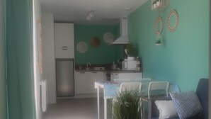 Une cuisine moderne avec des meubles blancs, un réfrigérateur et un coin repas comprenant une table et des chaises blanches. On y trouve un canapé bleu avec un coussin rayé et un coussin blanc. Une plante en pot est posée sur la table, et des assiettes décoratives sont accrochées au mur.