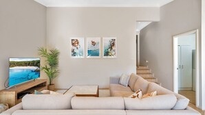 A living room with a beige sofa, a coffee table, a TV on a wooden stand, and three framed pictures on the wall.