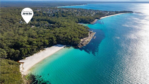 An aerial view of a coastal area with a beach, dense forest, and clear turquoise waters.
