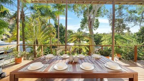 A wooden dining table set for six with white plates and silverware, surrounded by lush greenery and trees.