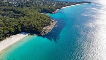 An aerial view of a coastal area with a beach, dense forest, and clear turquoise waters.