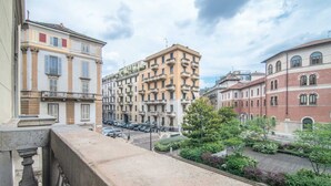 A balcony overlooking a street with multi-story buildings, a parked car, and a small garden area. - Sweett | Arco della Pace (Milano)