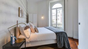A bedroom with a bed, a bedside table, a lamp, a window, and a view of buildings outside. - Sweett | Arco della Pace (Milano)