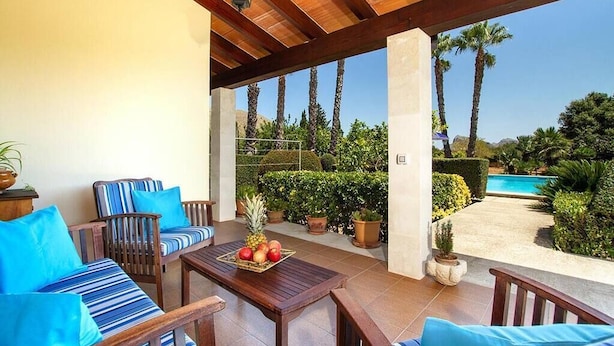 An outdoor patio area with wooden furniture, a table with fruit, and a view of a swimming pool and palm trees. - Villa Pantina, 2 bedroom villa near Pollensa (Port de Pollença)
