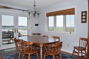 A dining room with a round wooden table and chairs, large windows with blinds, a chandelier, and a view of the outdoors.