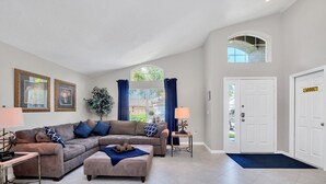 Ein Wohnzimmer mit einem Ecksofa, einem Couchtisch und einer Lampe. Es gibt eine Tür mit einem Schild, ein Fenster mit Vorhängen und eine Pflanze.