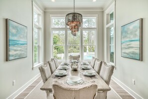 A dining room with a long table set for six, large windows, and two framed artworks on the walls.