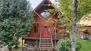 A log cabin with a red door, a wooden staircase leading to a porch, and a stone wall in the background.