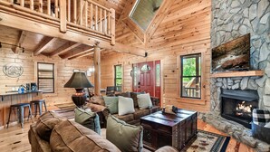 A cabin interior with a stone fireplace, wooden ceiling beams, and a cozy seating area.