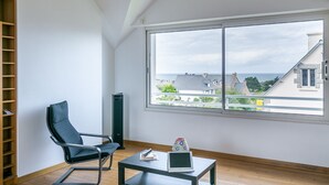 Une chambre moderne avec une grande fenêtre, une chaise noire, une petite table avec un ordinateur portable et des livres, et une bibliothèque.