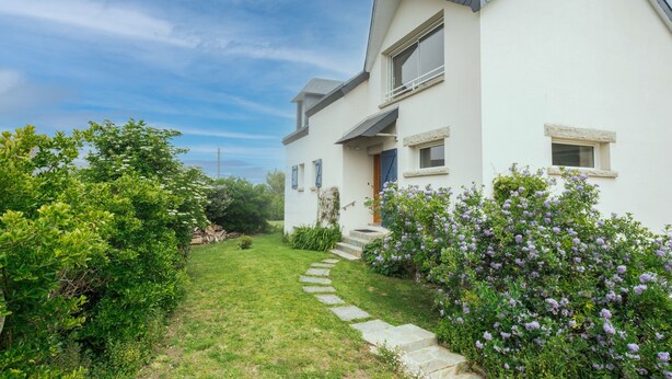 Une maison moderne à deux étages, avec un chemin de pierre menant à la porte d’entrée, entourée d’une végétation luxuriante et de fleurs violettes.