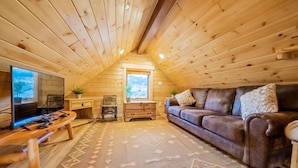 A cozy attic room with wooden walls and a sloped ceiling. It features a brown leather couch, a wooden coffee table, a flat-screen TV, and a patterned rug. - Elk Hotspot w/ Views, Deck & Game Room (Estes Park)