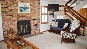 Ein Wohnzimmer mit Kamin aus Backstein, einer Couch, einem Korbstuhl, einem Holztisch und einer Treppe.