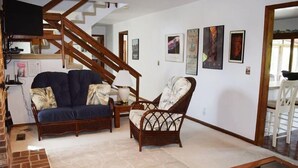 Ein Wohnzimmer mit einer Couch, einem Sessel und einem Glastisch. Im Hintergrund ist eine Treppe zu sehen.