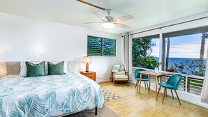 Une chambre à coucher comprenant un lit, une table de chevet, une chaise, une table, un ventilateur de plafond et une grande fenêtre donnant sur l’océan.