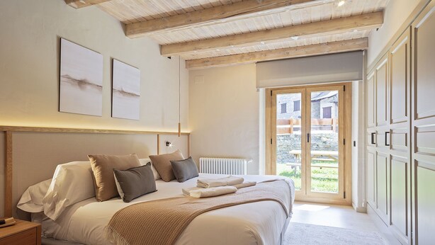 A bedroom with a large bed, a wooden ceiling, a radiator, and a view of a courtyard through a glass door. - Luderna - Apartamento con jardín Crabera (Arties)