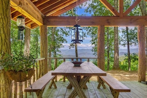 A wooden deck with a picnic table and hanging lanterns, surrounded by trees and a view of mountains.