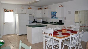 Une cuisine compacte avec des meubles blancs, un îlot central et un coin repas avec des chaises blanches et des sets de table rouges.