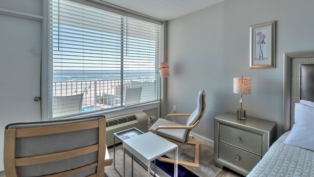 Une chambre d’hôtel comprenant un lit, un bureau, une chaise, une table de chevet, une lampe et une fenêtre avec des stores.