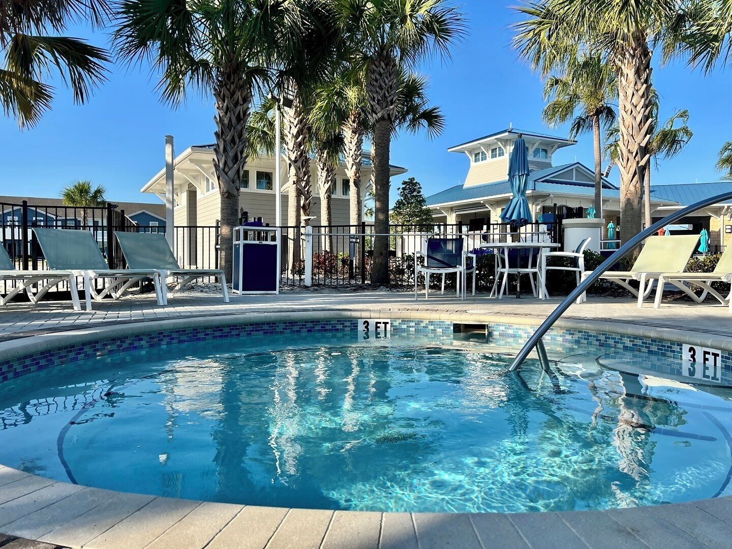 A clear swimming pool with lounge chairs and umbrellas, surrounded by palm trees and buildings.