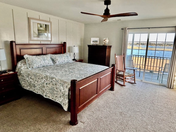 A bedroom with a wooden bed, a nightstand, a chair, a ceiling fan, a window with a view of a body of water, and a framed picture on the wall.