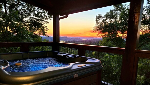 Ein Whirlpool auf einer Holzterrasse mit Blick auf einen Sonnenuntergang über einer bewaldeten Landschaft.
