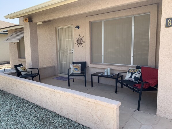 A covered patio area with a seating arrangement including two chairs and a table, a door with a decorative keyhole, and a large window with a screen.