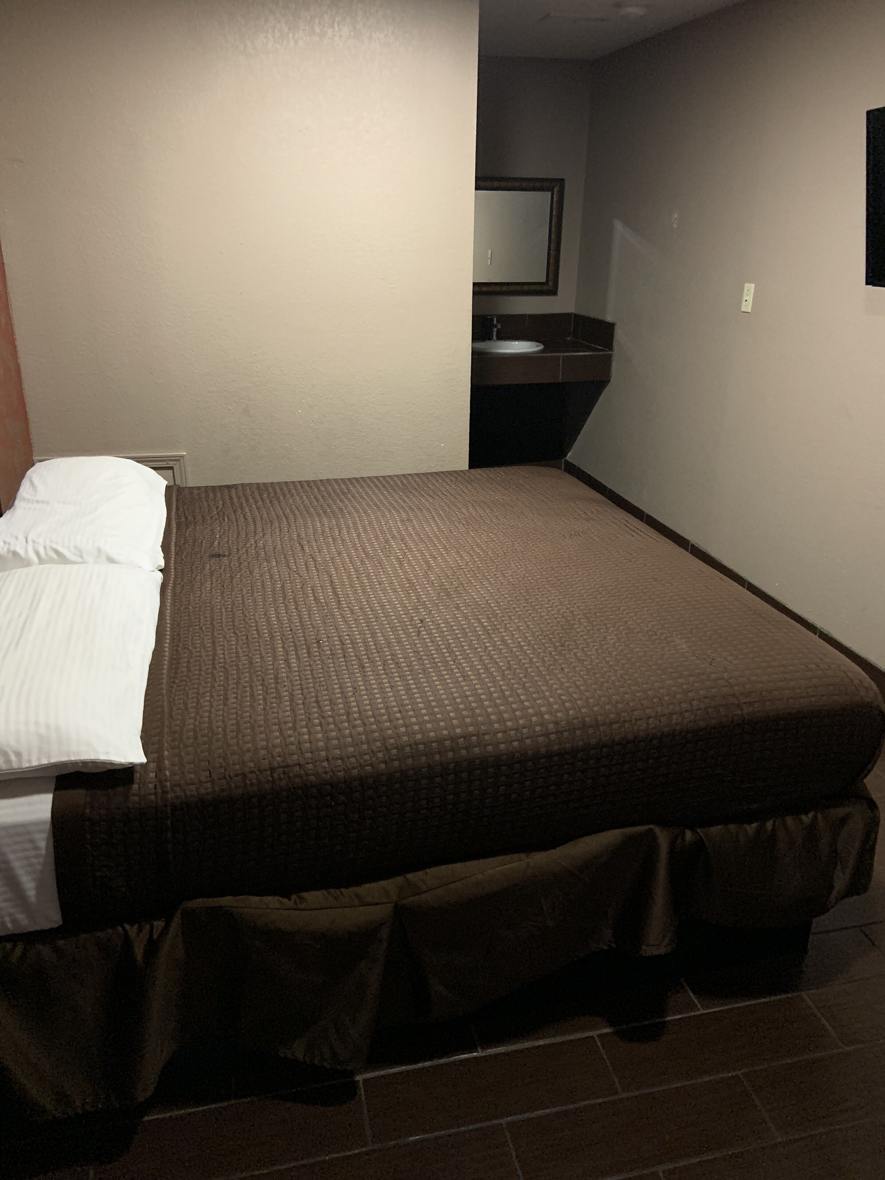 A small, neatly made bed with a brown cover, a white pillow, and a dark brown skirt. There is a small bathroom with a mirror and a sink in the corner.