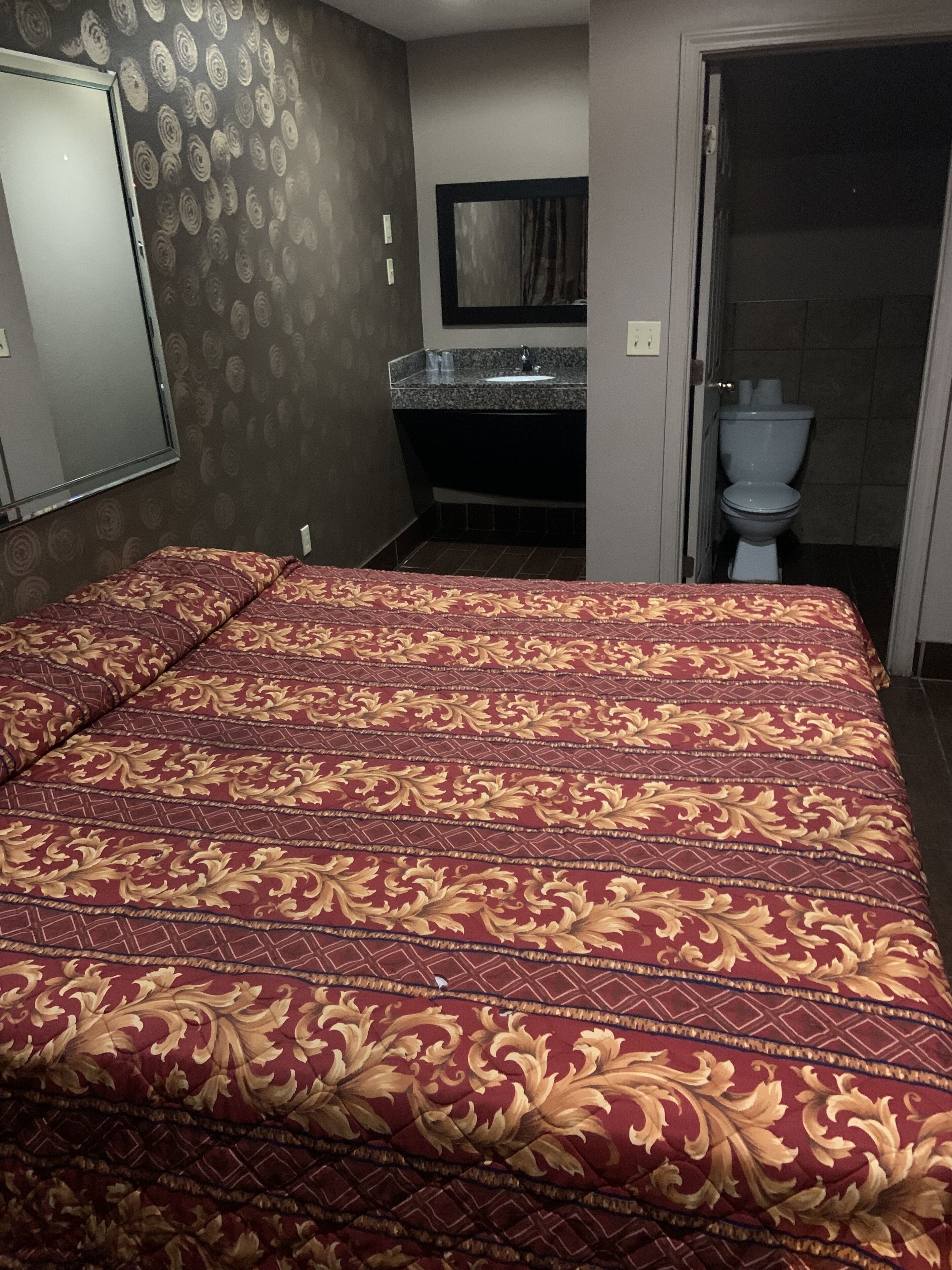 A room with a large bed, a bathroom with a toilet and sink, and a mirror on the wall.
