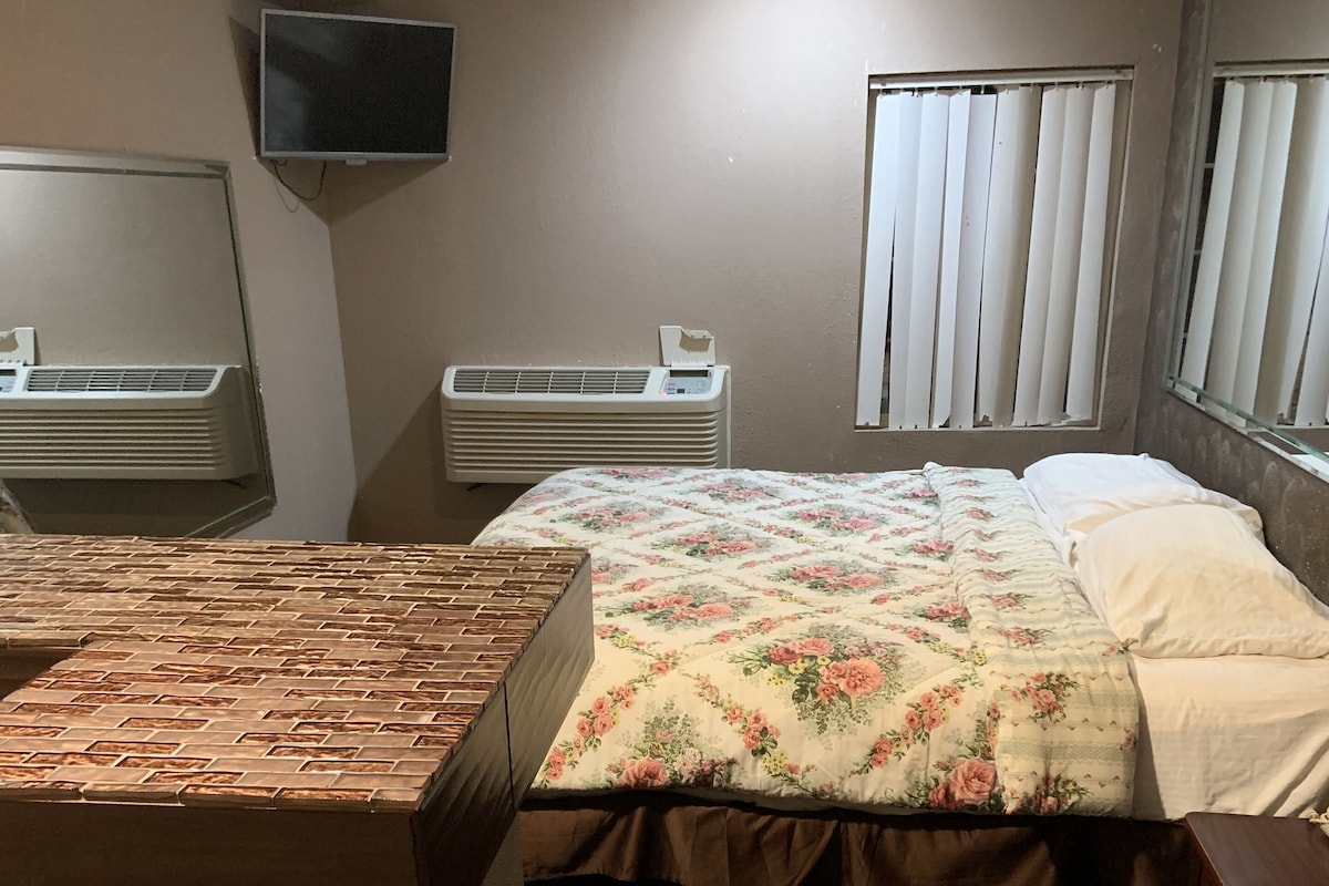 A bedroom with a bed, a TV mounted on the wall, an air conditioning unit, and a window with blinds.