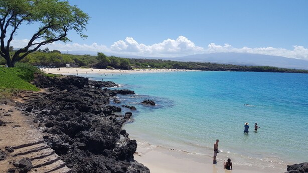 A beach with clear turquoise water, rocky shoreline, and a distant mountain range.