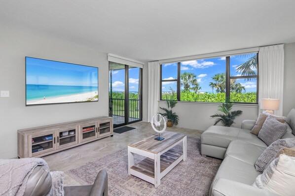 A living room with a large TV, a sofa, a coffee table, and a view of the beach through large windows.