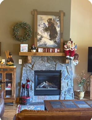 A cozy living room with a stone fireplace, a Christmas wreath, a painting of a skier, and a shelf with holiday decorations.