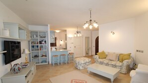 Ein Wohnzimmer mit einer Couch, einem Hocker, einem Couchtisch und einem Fernseher. Es gibt eine Küchenzeile mit einer Arbeitsfläche und Barhocker.