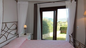 Ein Schlafzimmer mit Bett, Nachttisch, Lampe und Blick auf einen Garten und Hügel.