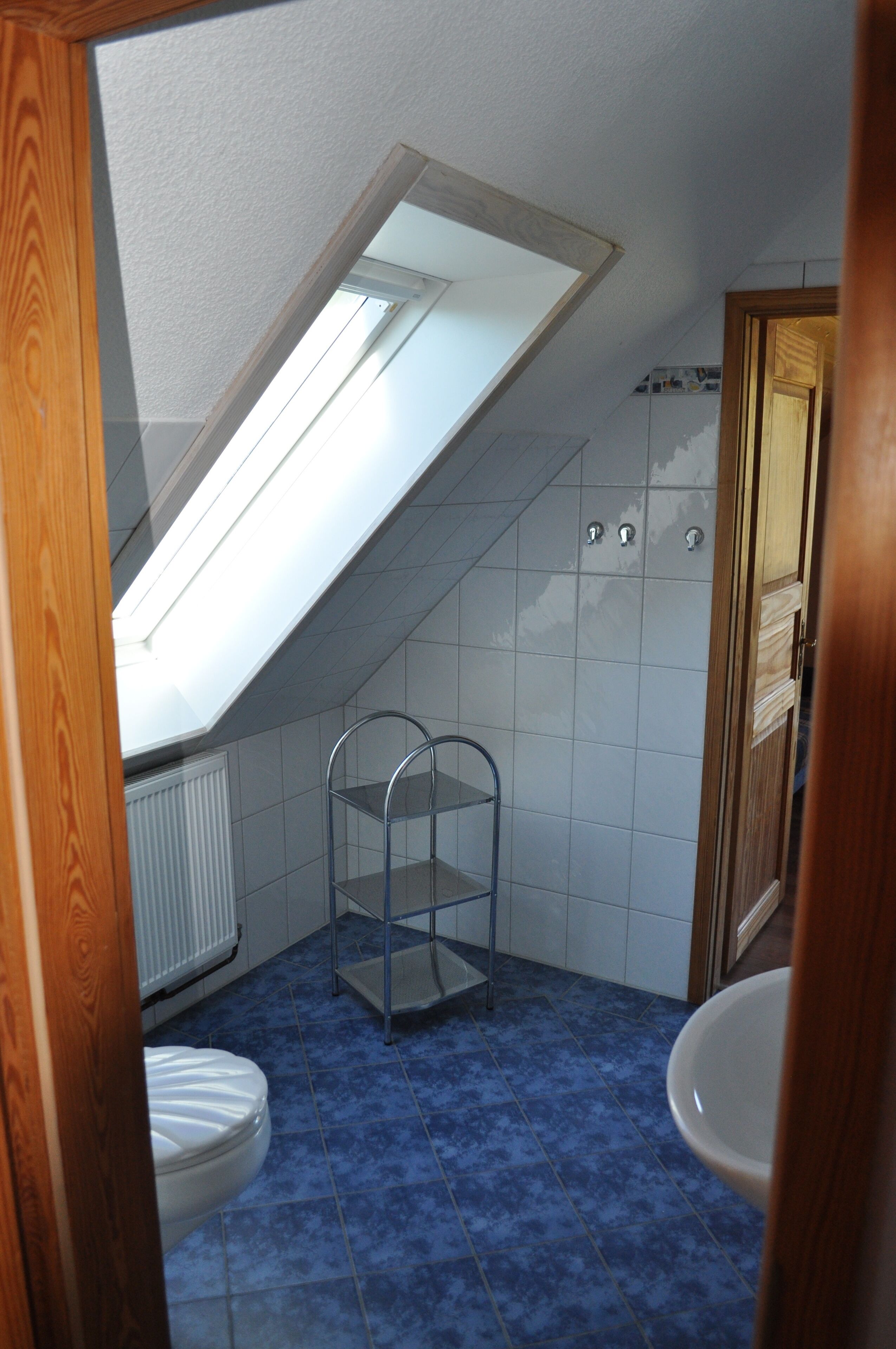 Ein kleines Badezimmer mit geneigtem Dach, einem Oberlicht, einem weißen Heizkörper, einem Metallregal und einer weißen Toilette.