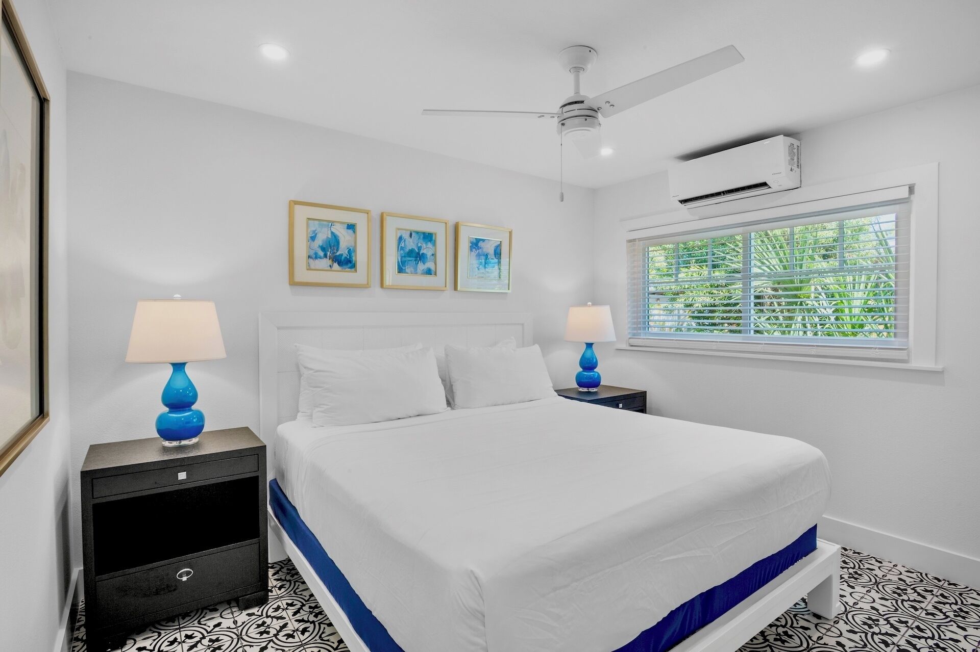 Quarto com cama, duas mesas de cabeceira, ventilador de teto, ar condicionado e janela com persianas.
