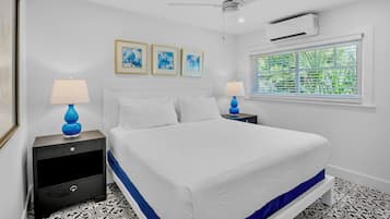 Quarto com cama, duas mesas de cabeceira, ventilador de teto, ar condicionado e janela com persianas.