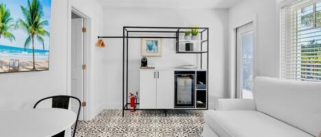En moderne stue med en hvid sofa, et spisebord med stole, et køkken med køleskab og skabe, samt et stort maleri med strandmotiv på væggen.