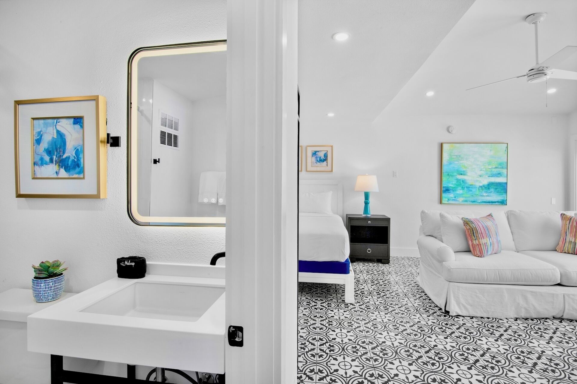 A modern hotel room with a white sink, a mirror, a bed with a blue mattress, a white sofa, and a patterned floor.