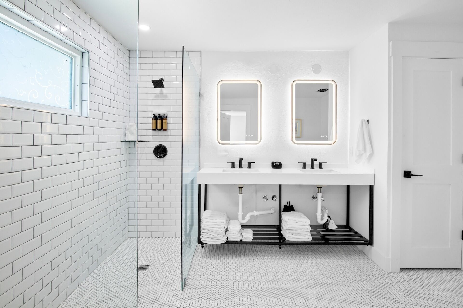 A modern bathroom with white tiled walls, a glass shower enclosure, a white sink with dual mirrors, and a towel rack with neatly folded towels.
